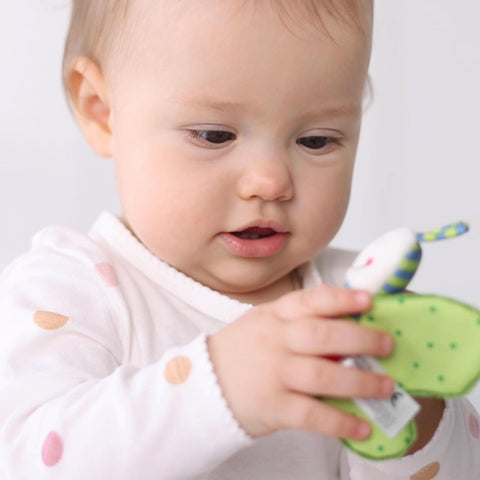 Das Baby im weißen Strampler mit pastellfarbenen Tupfen erkundet den grünen Knister-Schmetterling aus der Red Stars Collection und fördert so die Sinneswahrnehmung.