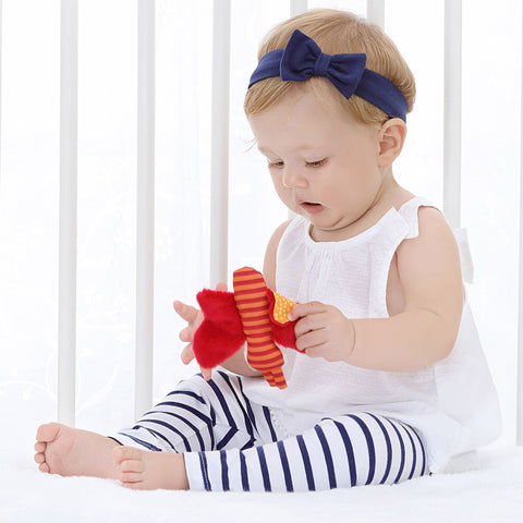 Ein Baby, das ein marineblaues Stirnband, ein weißes Oberteil und gestreifte Leggings trägt, sitzt auf dem Boden und hält einen orangefarbenen Knisterschmetterling aus Plüsch von Redstars.