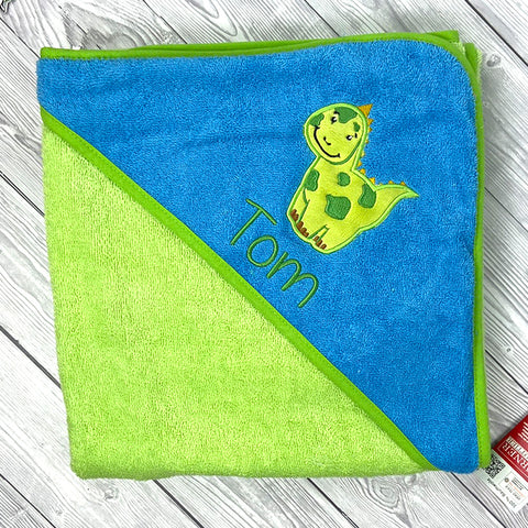 Ein gefaltetes Kapuzenbadetuch Limone/Ozean 100x100cm aus 100% Baumwolle mit Dinosaurier-Motiv und auf Wunsch mit Namen "Tom" bestickbar liegt auf einer hellen Holzoberfläche.