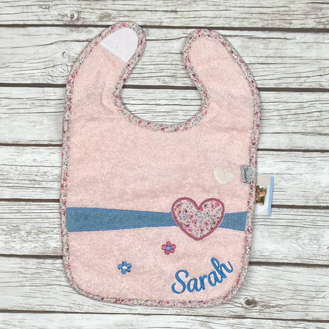Ein Plastik-Klettlätzchen Mabel Herz in Rosa mit Blumenbesatz und blauen Herzchen, personalisiert mit dem in Blau gestickten Namen Sarah unten rechts, wird auf einer Holzfläche gezeigt.