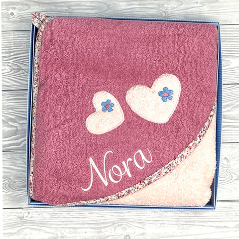 Ein rosafarbenes Kapuzenbadetuch Mabel Herz 100x100cm mit Blumenbesatz, zwei Herzverzierungen und dem Namen Nora personalisierbar gestickt, ausgestellt in einer Box auf einer Holzfläche.