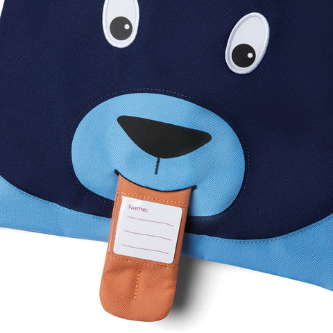 Eine Nahaufnahme des blauen Affenzahn Turnbeutels - Bär aus recyceltem Polyester mit Cartoon-Augen, einer Nase und einem orangefarbenen, zungenförmigen Anhänger, der mit einem Namensetikett personalisiert werden kann.