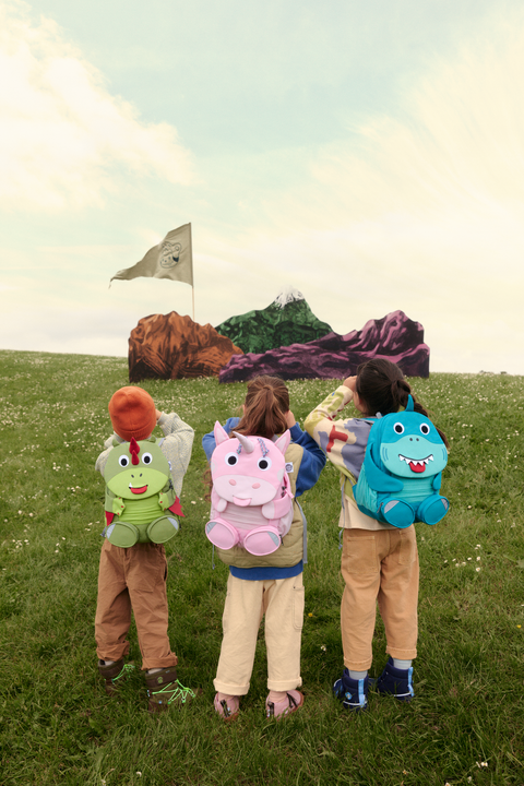 „Drei Kinder mit bunten Tier-Rucksäcken (Drache, Einhorn, Hai) stehen auf einer Wiese und blicken auf ein gemaltes Bergpanorama mit Fahne.