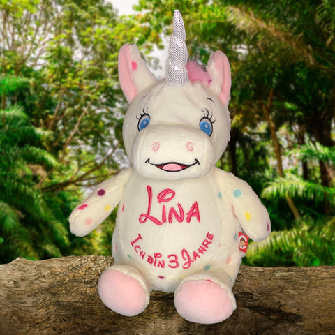 Aurora, das gepunktete Einhorn, ein weißes Plüschtier-Geschenk mit silbernem Horn, bunten Flecken und blauen Augen, sitzt auf einem Baumstamm. Die persönliche Stickerei "Lina Ich bin 39 Jahre" ziert die Front zwischen grünen Bäumen.
