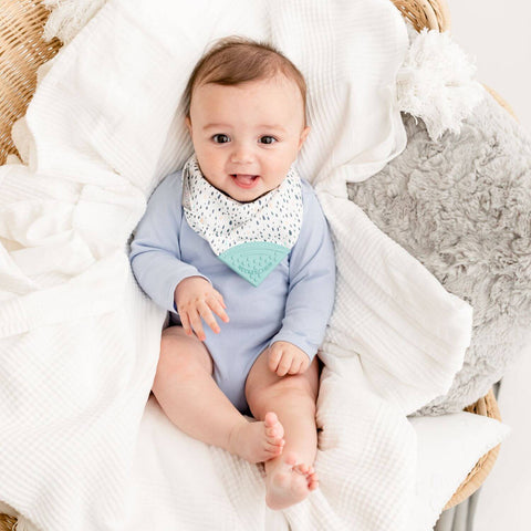 Ein Baby in einem hellblauen Strampler und einem Neckerchew Zahnen Dribble Lätzchen - Birnendrops sitzt auf einer weißen Decke in einem Weidenkorb und lächelt in die Kamera.