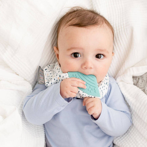 Ein Baby im hellblauen Hemd liegt auf einer weißen Decke und kaut auf dem Neckerchew Zahnen Dribble Lätzchen - Birnentropfen Beißlätzchen.