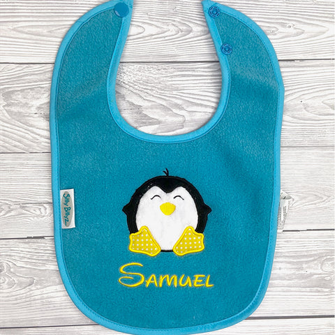 Aqua Silly Billyz Lätzchen, Vlies: Blaues, bleifreies & PVC-freies Baby-Lätzchen mit lachendem Pinguin und gelbem "Samuel"-Schriftzug unter dem Pinguin.