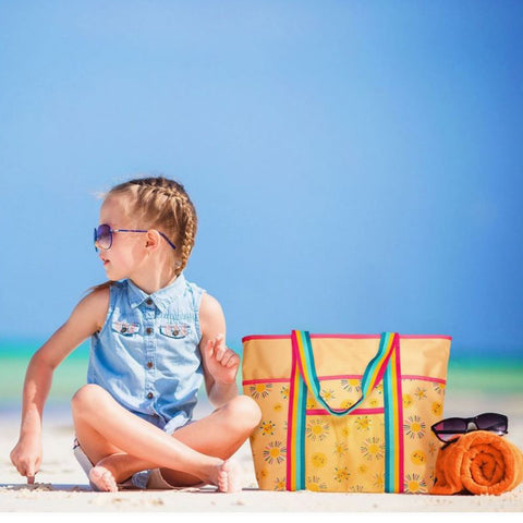 Ein junges Mädchen mit Sonnenbrille und ärmellosem Oberteil sitzt mit ihrer Großen Strandtasche - Dino mit fröhlichen Dinosaurier-Motiven - an einem Sandstrand, neben einem orangefarbenen Handtuch und einer Sonnenbrille, das Meer und der blaue Himmel im Hintergrund.