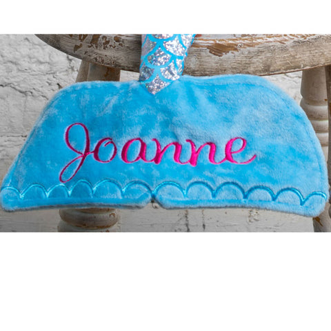 Eine blaue Meerjungfrau Stoffpuppe aus hochwertigem Material mit dem Namen "Joanne" in pinker Stickerei, sitzt auf einem Holzstuhl.