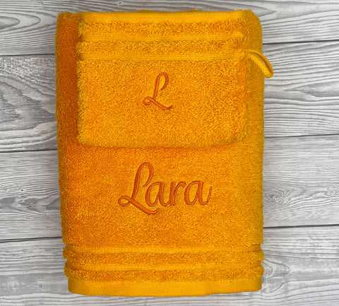 Ein gefaltetes Handtuch Calypso Feeling, Sonne 50x100cm plus Waschhandschuh ruht auf Holz, mit "Lara" und dem Initial "L" in orange bestickt - eine elegante Möglichkeit, Ihr Badetuch zu personalisieren.