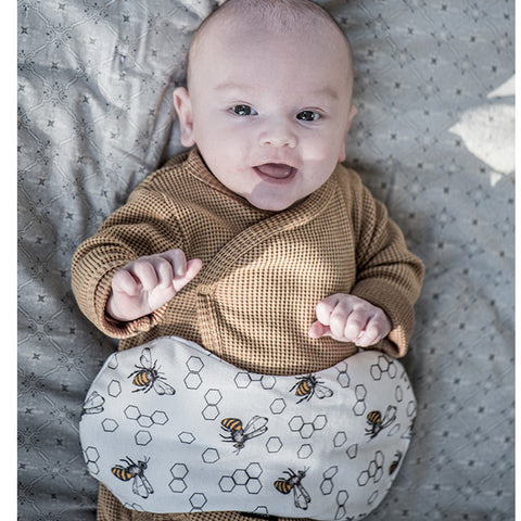 Ein Baby in einem braunen Outfit liegt auf einer gemusterten Decke und hat ein KipKep Wärmekissen - ECO Biene Special mit Bienen und Sechsecken über der Taille.