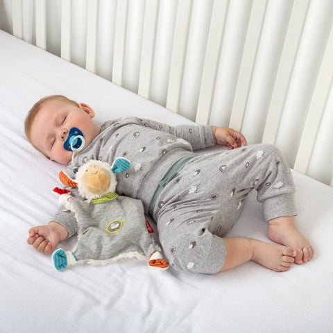 Ein Baby im grauen Schlafanzug schläft auf einer weißen Matratze, kuschelt mit dem Schnuffeltuch Schaf Boller Schäfle und einem blauen Schnuller, daneben liegt ein zartes Kuscheltier.