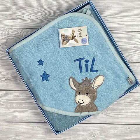 Das Kapuzenbadetuch Esel Emmi in Mittelblau mit Sternen (100x100cm) ist ordentlich gefaltet in einer Geschenkbox auf einer Holzoberfläche platziert.