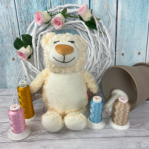 Der Wächter Bär mit Flügeln sitzt vor einem weißen Kranz mit rosa Rosen, umgeben von gelben, rosa, blauen und beigen Fadenspulen und einem umgekippten beigen Topf auf einer Holzfläche.