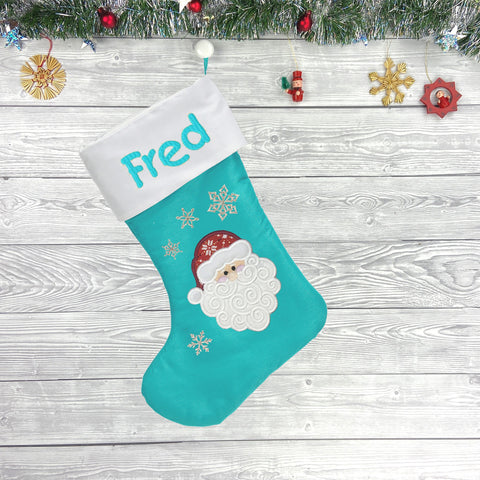 Ein eisblauer Weihnachtsstrumpf personalisiert (Stickerei im Preis inklus) mit Fred in Buchstaben, einem Santa-Gesicht und Schneeflocken hängt vor Holz-Hintergrund mit festlicher Deko.