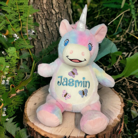 Starflower, das Cubbie Einhorn, ein pastellfarbenes personalisiertes Plüschtier mit Jasmin- und Schmetterlingsdesign, sitzt auf einem Baumstumpf zwischen grünen Pflanzen-ein süßes Geschenk zum Geburtstag.