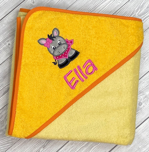 Das Kapuzenbadetuch Hellgelb/Gold 100x100cm ist ein gelbes Kinderhandtuch aus Baumwolle mit orangefarbenem Rand. Auf der Kapuze ist ein Cartoon-Esel mit Schleife und der Name Ella in rosa aufgestickt.
