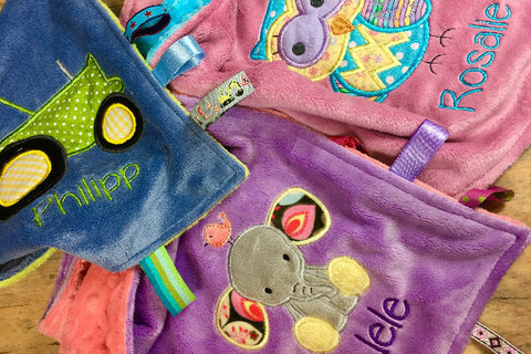Die Schmusekind Ersatz-Decke: Ihre Schmusedecke neu gefertigt besteht aus farbenfrohen Mikrofaser-Babydecken mit Tiermotiven und Namen, wie zum Beispiel einer Eule namens Rosalie und einem Elefanten namens Melele, die auf einer hölzernen Oberfläche angebracht sind.