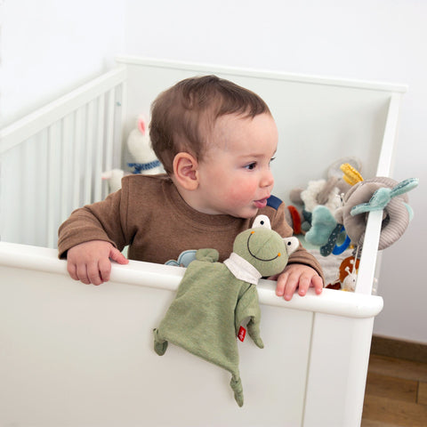 Ein Baby steht in einem weißen Kinderbettchen und hält ein Mini Jersey Schnuffeltuch Frosch aus Bio-Baumwolljersey in der Hand, umgeben von anderen Stofftieren, die die sensorische Entwicklung unterstützen.