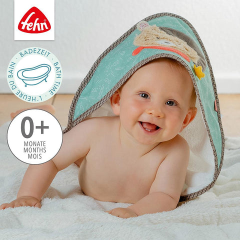 Ein lächelndes Baby liegt auf einer weißen Decke, teilweise bedeckt mit dem mintgrünen Kapuzenbadetuch Bär. Dieses Kapuzenbadetuch von Frottee ist mit niedlichen Tiermotiven versehen und für das Alter 0+ Monate geeignet, wie durch Symbole und Text angegeben.