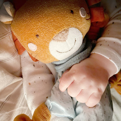 Eine Babyhand hält das weiche Baby Schnuffeltuch Löwe, während es auf einem gemusterten Stoff ruht - ideal zum Kuscheln und Bequemlichkeit.
