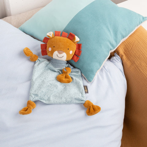 Ein Baby Schnuffeltuch Löwe ruht auf einem ordentlich gemachten Bett mit hellblauer Bettwäsche, einem blaugrünen Kissen und einer senffarbenen Decke und lädt die Kleinen zum Kuscheln ein.
