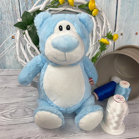 Der maschinenwaschbare "Blauer Bär - Cubbyford" sitzt auf einer Holzfläche neben Garnspulen, einem Blumenkranz und einem umgestürzten Behälter im Hintergrund - das perfekte Geschenk!.